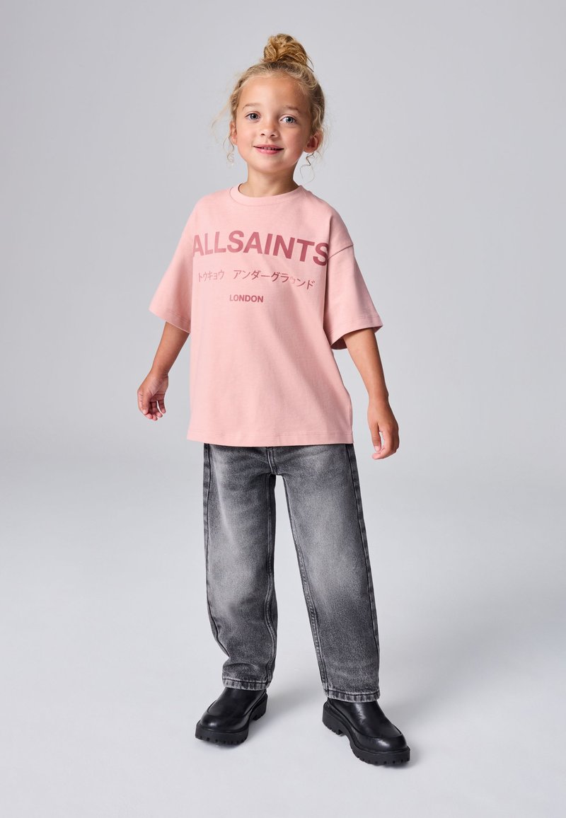 Vaaleanpunainen yläosan kokoinen t-paita, jossa on tummemman sävyn "ALLSAINTS"-logo, yhdistetty haalistuneisiin harmaisiin leveälahkeisiin farkkuihin ja paksupohjaisiin mustiin kenkiin.