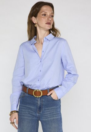 Donna con camicia azzurra chiusa con bottoni infilata nei jeans blu con cintura marrone, che indossa orecchini a cerchio dorati e braccialetti, con la mano in tasca.