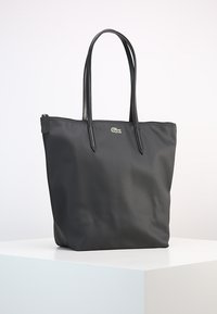 Borsa tote nera con superficie texturizzata, due manici lunghi, chiusura con zip e un piccolo logo Lacoste davanti. Design semplice e funzionale.