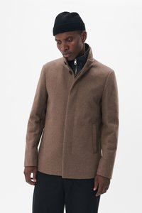 Abrigo de mezcla de lana marrón con cuello alto, con un tapeto oculto, bolsillos laterales y un corte entallado. El modelo lleva un gorro de punto negro.