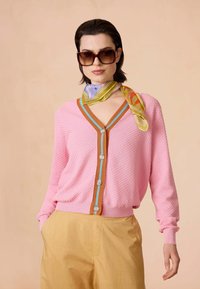 Roze textuur cardigan met een V-hals, oranje en blauwe afwerking, en zes knopen. Gecombineerd met beige broek en een multikleurige zijden sjaal.