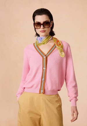 Roze textuur cardigan met een V-hals, oranje en blauwe afwerking, en zes knopen. Gecombineerd met beige broek en een multikleurige zijden sjaal.