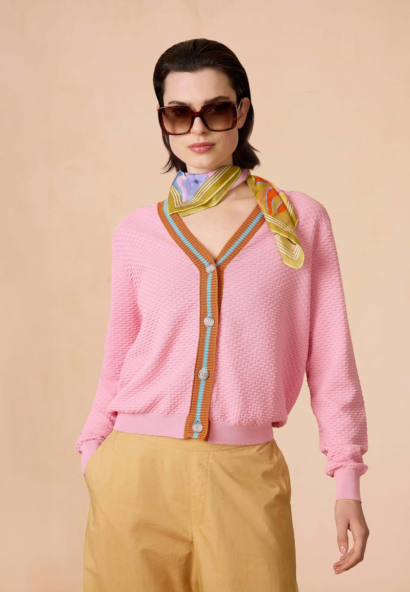 Roze textuur cardigan met een V-hals, oranje en blauwe afwerking, en zes knopen. Gecombineerd met beige broek en een multikleurige zijden sjaal.