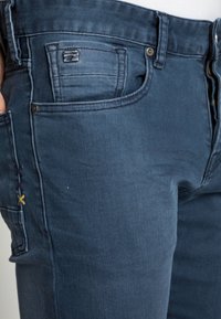 Acercamiento a unos jeans de mezclilla azul oscuro con bolsillos frontales y laterales visibles, detalle de costura en cruz amarilla y remaches de metal.