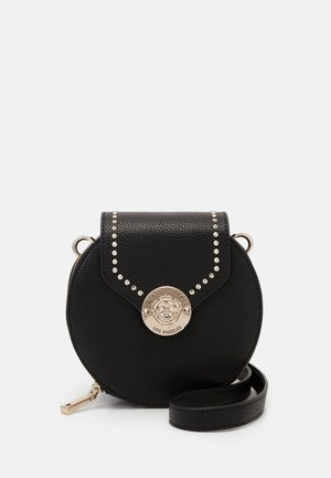 Zwarte ronde lederen crossbodytas met zilverkleurige studs, voorflap en ronde gouden logoplaat met de tekst "Guess 1981 Los Angeles."