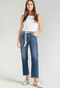 Jean en denim bleu clair à taille haute avec des coutures apparentes, des ourlets effilochés et une ceinture à cordon style corde. Associé à un débardeur blanc.