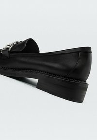 Sorte læderloafer med lav stakhed heel og sølv kædeddetail på vamsen mod en hvid baggrund.