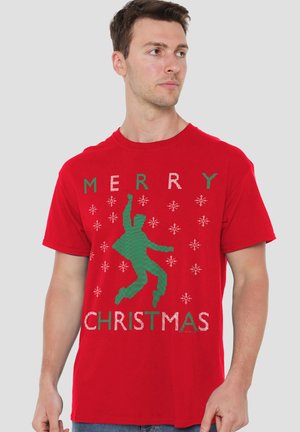 Mann trägt rotes T-Shirt mit grünem Umriss einer tanzenden Person und dem Text "Frohe Weihnachten", umgeben von weißen Schneeflocken.