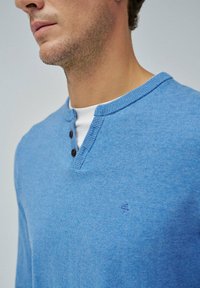 Maglione a maglia azzurro chiaro con colletto henley, dotato di due bottoni scuri e un piccolo logo sul petto. Texture morbida, design informale.