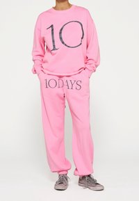 Sudadera rosa con gran estampado negro "10", combinada con pantalones deportivos a juego que llevan "10DAYS" en negro. El tejido es suave; los pantalones tienen puños elásticos.