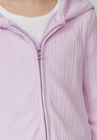 Helles lila Zip-Hoodie mit vertikaler Rippstruktur, mit passendem Reißverschluss und einer weichen Kapuze. Darunter ist ein weißes Shirt sichtbar.