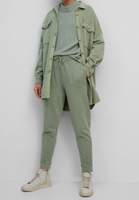Persoon in een saliegroen gebreid sweater, bijpassende joggerbroek, oversized knopenjack en off-white high-top sneakers.