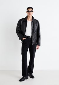 Zwarte leren jas met rits, witte getailleerde t-shirt, zwarte denim jeans en zwarte strakke schoenen; model met kort haar die donkere zonnebril draagt.