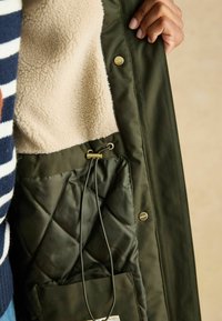 Zielona kurtka z zewnętrzną warstwą, beżowym polarem wewnątrz, pikowaną podszewką, ściągaczem i złotymi akcentami.