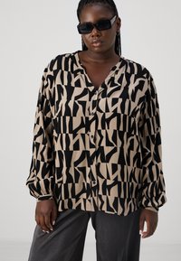 Chemise à boutons noire et beige à motif, avec des manches longues, en texture douce, présentant des formes géométriques irrégulières et un décolleté en V.