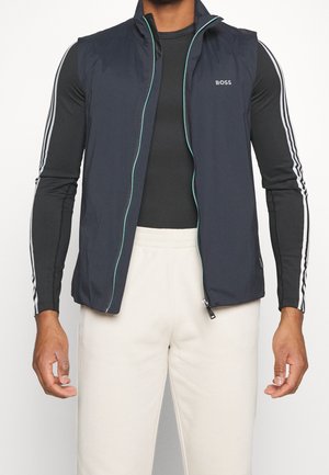Mann trägt ein schwarzes Langarmshirt mit weißen Streifen und eine marineblaue ärmellose Weste mit Reißverschluss, kombiniert mit weißen Hosen.