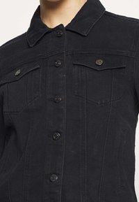 Svart jeansjacka med krage, framknäppning med knappar, två bröstfickor och vertikala sömdetaljer. Slät textur med metalliska accenter.