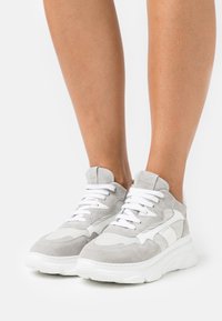 Jambes portant des baskets en daim gris clair et blanc avec des semelles blanches épaisses, debout sur une surface blanche.