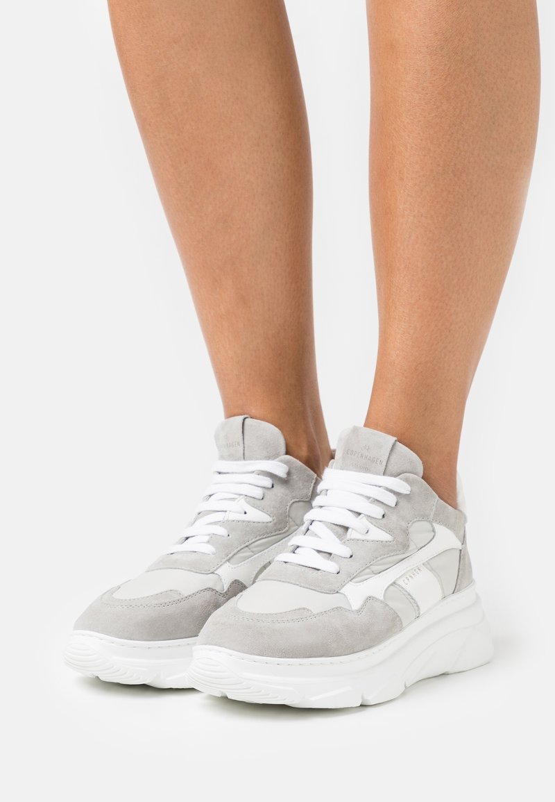 Jambes portant des baskets en daim gris clair et blanc avec des semelles blanches épaisses, debout sur une surface blanche.