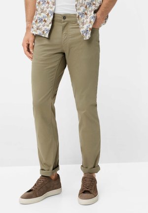 Homme portant un pantalon olive retroussé, des baskets en daim marron et une chemise à fleurs, les mains dans les poches, debout devant un fond blanc.
