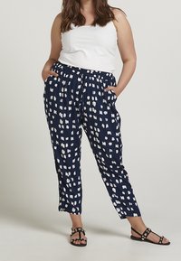 Pantaloni blu con motivo a chiazze bianche e astratte, vita elastica, tasche laterali, gamba affusolata, abbinati a un top bianco senza maniche e sandali neri.