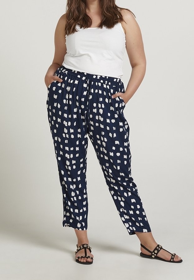 Pantaloni blu con motivo a chiazze bianche e astratte, vita elastica, tasche laterali, gamba affusolata, abbinati a un top bianco senza maniche e sandali neri.