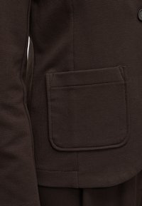 Veste en tissu brun texturé avec une poche plaquée rectangulaire à l'avant et un petit bouton en métal près du bord supérieur.