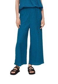 Vrouw in losse blauwe textuur broek met een verstelbare tailleband en zwarte sandalen met dubbele bandjes, staande tegen een witte achtergrond.