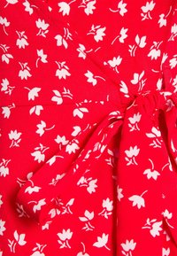 Roter Stoff mit einem Blumenmuster aus weißen Blumen. Das Material wirkt weich, mit einem Knoten-Detail an der Taille, das einen gerafften Look erzeugt.