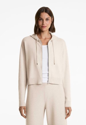 Woman wearing beige hooded zip-up cardigan over white tank top and matching beige pants, standing against white background.