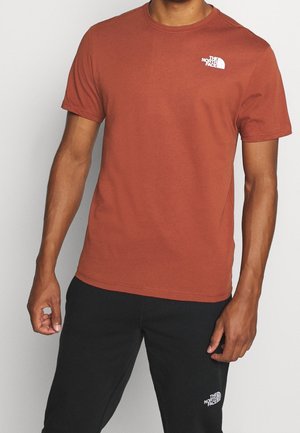 Hombre con camiseta de manga corta color óxido y pantalones negros, ambos con logos blancos de The North Face en el pecho y el muslo.