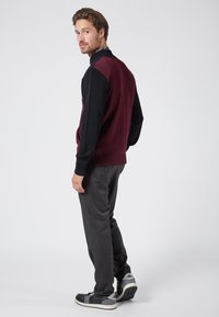 Dunkelrot und schwarze Strickjacke mit hohem Kragen, kombiniert mit grauen Stoffhosen und schwarzen Sneakers mit grauen Akzenten.