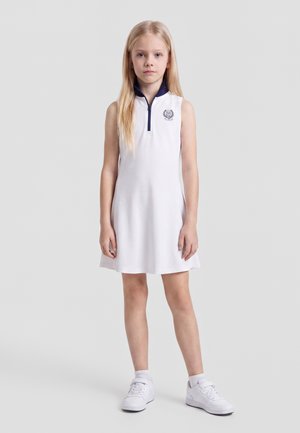 Jeune fille debout, face à l’avant, portant une robe blanche sans manches avec un col bleu marine et des baskets blanches sur un fond clair uni.