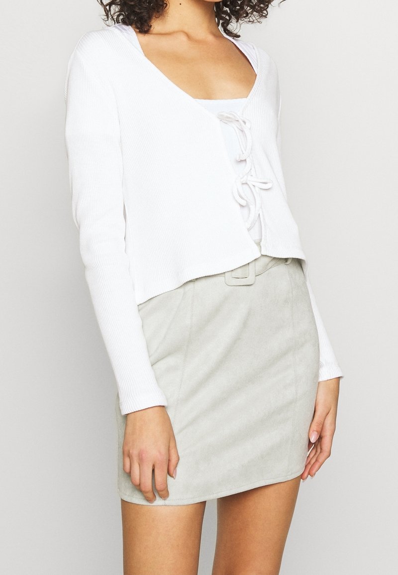 Cardigan court blanc côtelé avec manches longues et détail à lacets sur le devant, associé à une mini-jupe en daim gris clair avec une ceinture.