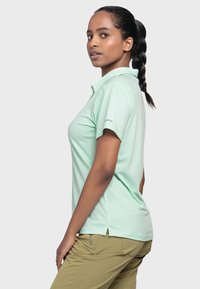 Mintgrünes Polohemd mit Kragen, kurzen Ärmeln und leichten Seitenschlitzen. Mit einem Logo auf dem linken Ärmel. Kombiniert mit Khakihosen.