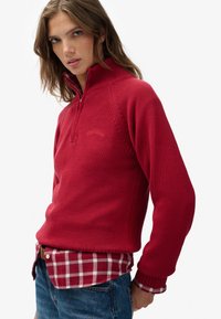 Czerwony dziergany pulower z wysokim kołnierzem i zapięciem na zamek do połowy długości. Posiada ściągacze na mankietach oraz teksturowany wzór, noszony na warstwie koszuli w kratę.