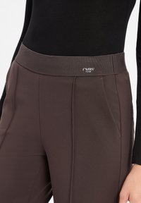 Pantalons marron en tissu extensible et lisse, avec une taille haute et des poches avant ; logo sur la taille.
