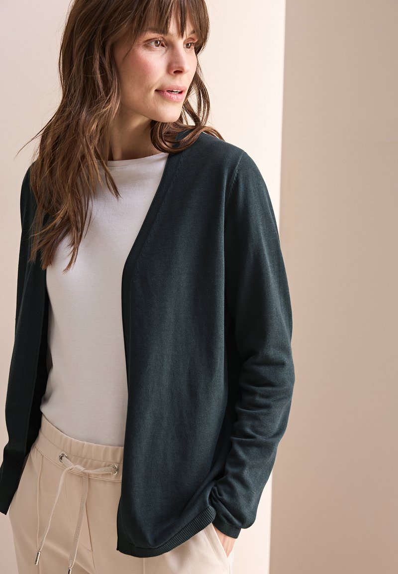 Mörkgrön cardigan i mjukt material, med v-ringad design och avslappnad passform. Buren över en ljusgrå skjorta och matchad med beige byxor.