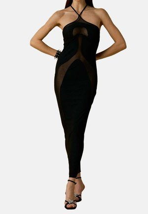 Vrouw draagt een mouwloze zwarte jurk met doorzichtige panelen, een gekruiste halterhalslijn en zwarte hoge hak sandalen met enkellinten.