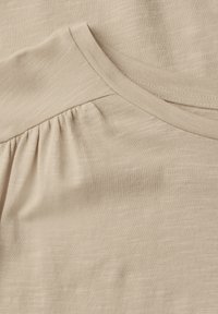 Beige pletená tkanina s kulatým výstřihem a nařasením na rameni, která vykazuje měkký, texturovaný povrch.