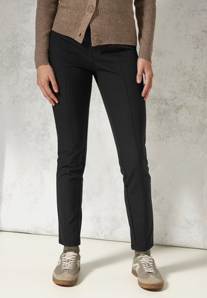 Černé kalhoty slim fit s hladkou texturou, dvěma předními kapsami a prošívanými detaily. V kombinaci s béžovými teniskami a hnědým kardiganem.