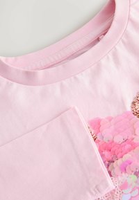 Helles pinkes T-Shirt mit rundem Ausschnitt, das eine Tasche und ein Herzdesign aus glänzenden pinken Pailletten aufweist.