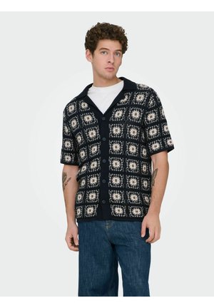 Junger Mann mit lockigem Haar, der ein schwarz-weiß gemustertes kurzärmeliges Hemd über einem weißen T-Shirt und blauen Jeans trägt.