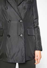 Veste noire à double boutonnage en tissu satin, avec de grands boutons noirs et deux poches avant, dotée d'un col cranté.