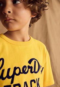Geel katoenen T-shirt met korte mouwen, voorzien van marineblauwe tekst "SuperDry TRACK" in een dikke, cursieve stijl. Ribgebreide ronde hals.