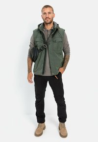 Grünes ärmelloses Oberteil mit mehreren Taschen, darunter ein graues Langarmhemd, schwarze Hose und beige Stiefel. Umhängetasche über der Schulter getragen.