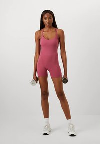 Rozā pieguļošs sporta kombinezons ar plānām lencītēm, ko valkā persona, turot hanteles. Balti sporta zeķes un apavi papildina izskatu.