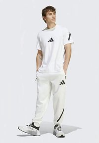 Balta Adidas T-krekls ar melnu logotipu, apvienots ar baltām sporta biksēm ar melniem svītrainiem un logotipa akcentiem. Melni un pelēki sporta apavi.