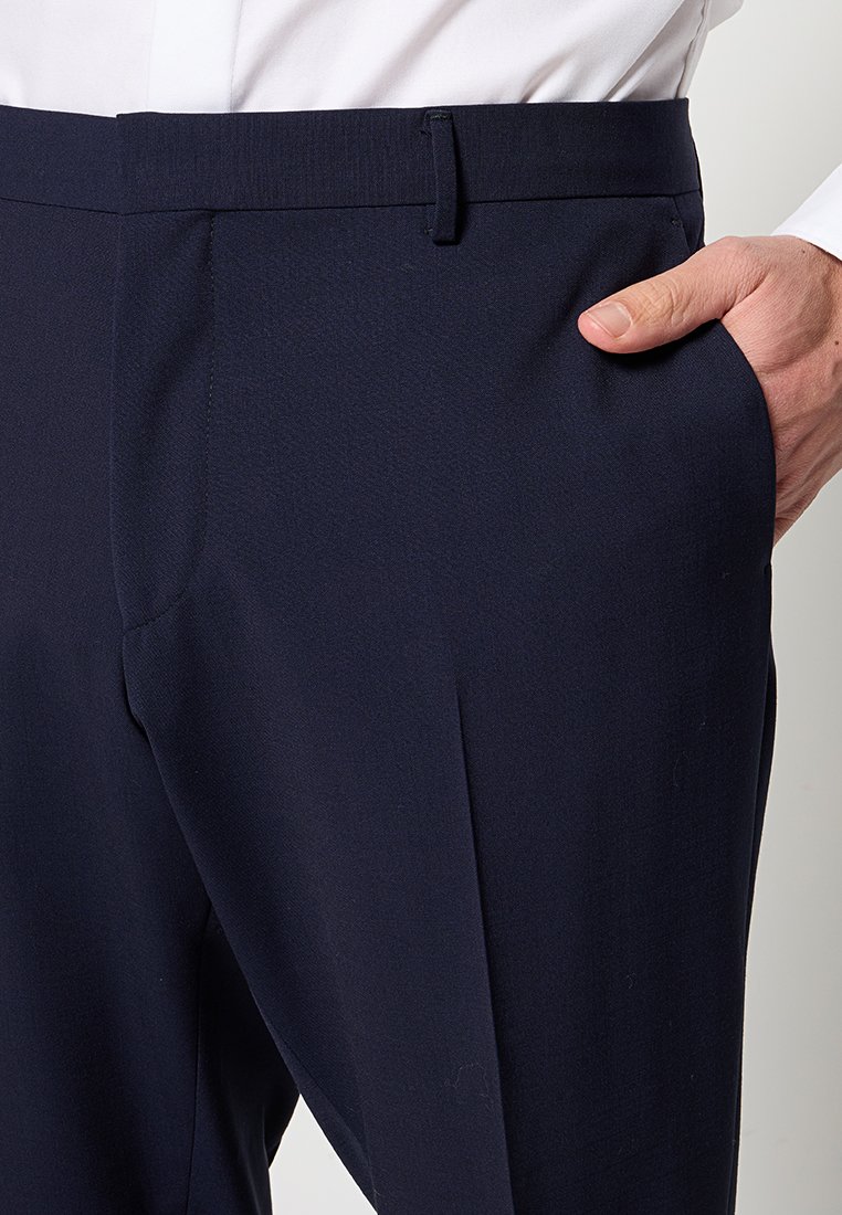 Mann in dunkelblauen Anzughosen mit der Hand in der rechten Tasche, kombiniert mit einem weißem Hemd, das in die Hose gesteckt ist.