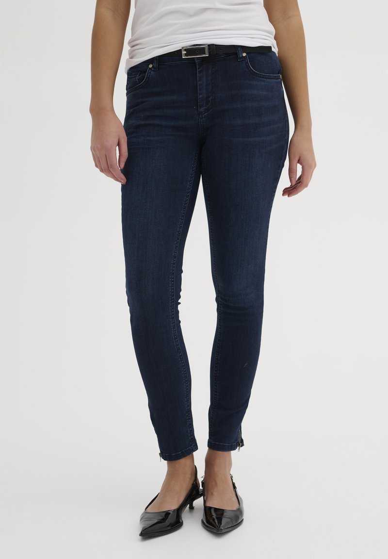 Temno modre skinny kavbojke iz denima. Značilnosti vključujejo zadrgo na gležnjih, dizajn s petimi žepi in ozko prileganje za eleganten videz.